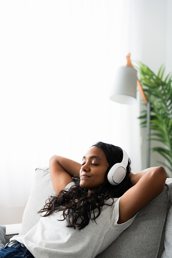 Young Indian woman relaxing sitting on sofa with copy space. Teenager listening to music on headphones.