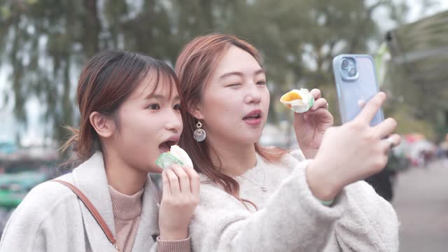 At Cheung Chang, Hong Kong, two young women happily enjoy steamed buns while capturing a selfie, both wearing smiles and relishing their food