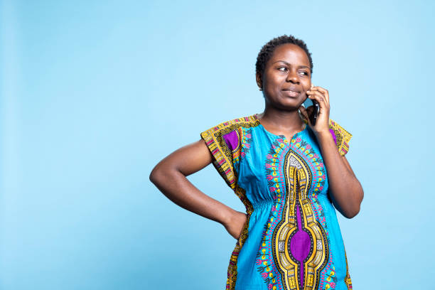 African american woman chatting on phone call in studio, answering telephone line to have remote conversation with her friends or family. Young person in traditional ethnic attire.