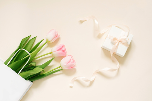 Flat lay composition with pink yellow tulips near a white paper bag with gift on a beige background with copy space. Valentin Day, Birthday, Mother day