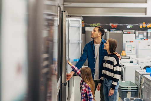 Cheerful parents and daughter choose a refrigerator for their new apartment in a wholesale store in the home appliance section