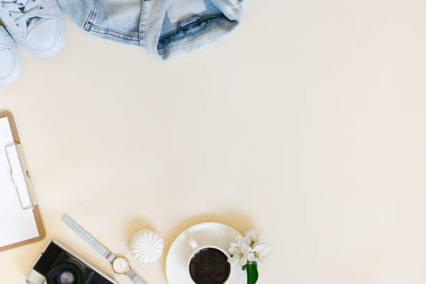 Planning a summer trip. A tablet with a blank sheet of paper, a retro camera, white apple blossoms, a watch, women's clothing and a cup of coffee on a beige background with a copy space. The workplace of a travel blogger