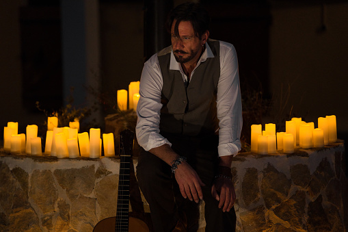Professional guitarist in waistcoat resting after playing Spanish guitar. The man is leaning against the street lamp in the town square, surrounded by candles.