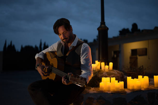 um guitarrista profissional de colete toca guitarra espanhola. o homem está encostado a um poste de iluminação pública na praça da cidade, cercado por velas ao entardecer. - plaza de espana sevilla town square seville - fotografias e filmes do acervo