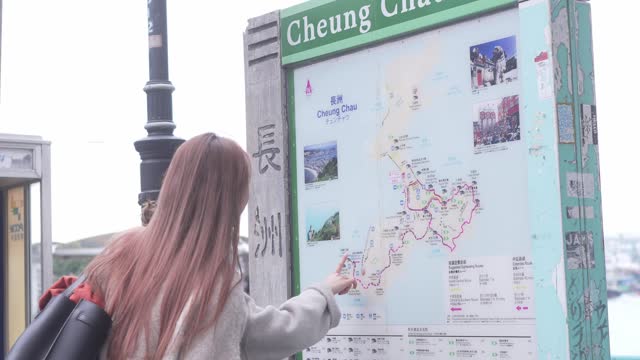 In Cheung Chang, Hong Kong, a woman with pink hair and a white jacket examines a map on a sign affixed to a post, featuring a green background,.