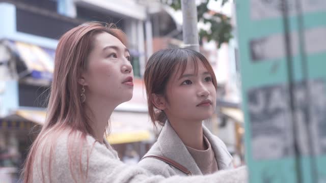 The scene in Cheung Chang, Hong Kong, captures two young women scrutinizing a poster on a wall. One woman displays long brown hair, while her companion exhibits long reddish-brown hair.