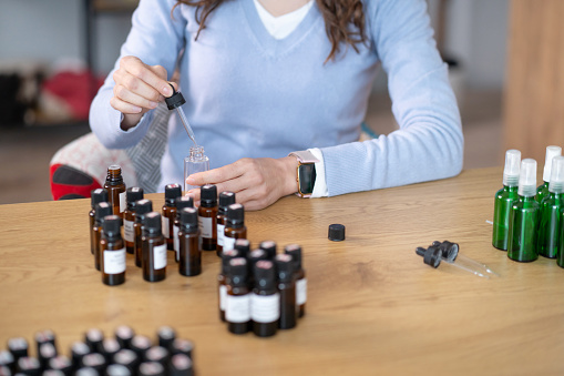 A woman is seated at a table surrounded by numerous bottles, engaged in the process of preparing Bach flower remedy. She is focused and actively working on mixing and diluting the tinctures in various containers.