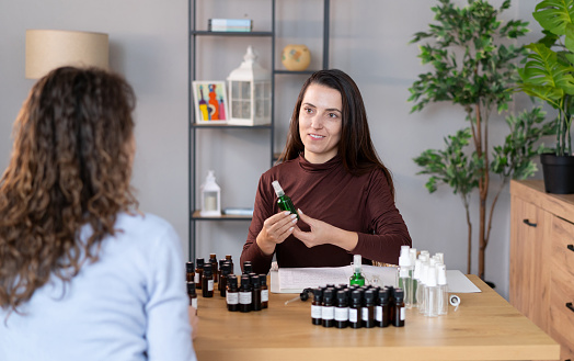 A woman is seated at a table, surrounded by various bottles of essential oils. She is focused on preparing a Bach flower remedy, carefully selecting and blending the oils for the mixture.