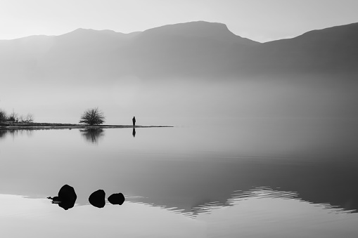 Black and white photo of magnificient landscape: a lake between high mountains. The water is reflecting the surroundings and the sky