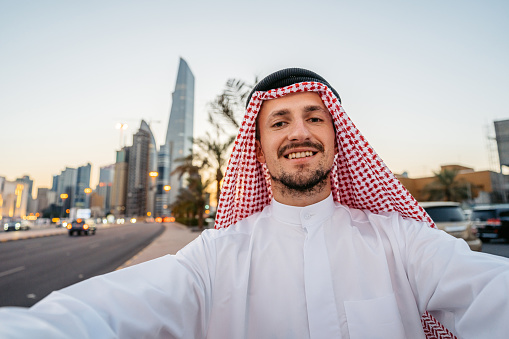 Handsome young man wearing keffiyeh and dishdasha taking selfies using his smart phone on the street in Kuwait city in Kuwait.