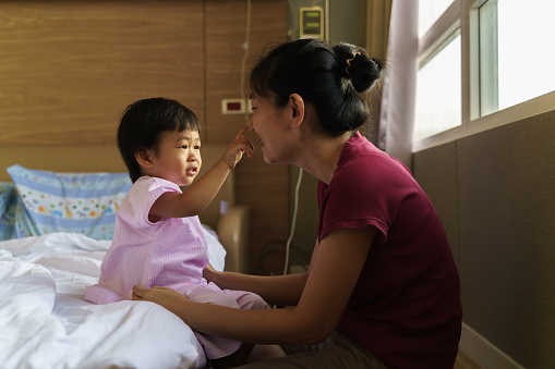 Asian little boy with Saline intravenous drip sitting in the hospital bed while mother holding his hand comforting. Adorable Asian kids get sick from virus and hospitalized. Baby health care medical concept.