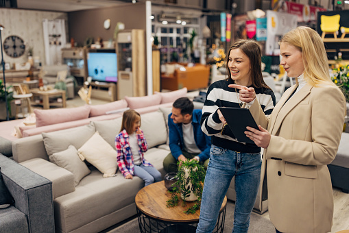 The saleswoman uses a digital tablet to show the cheerful female customer an offer of different sofa beds that they have in stock