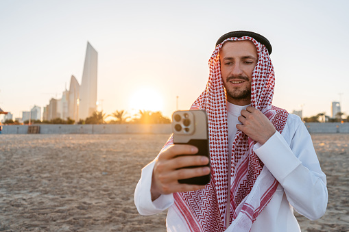 Handsome young man wearing keffiyeh and dishdasha taking selfies using his smart phone on the beach in Kuwait city in Kuwait.