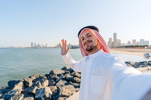 Handsome young man wearing keffiyeh and dishdasha having a video call using his smart phone on the beach in Kuwait city in Kuwait.