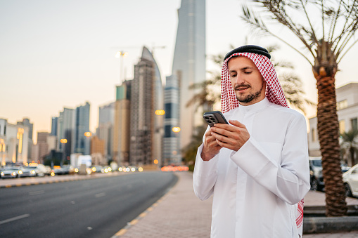 Handsome young man wearing keffiyeh and dishdasha and using his smart phone on the street in Kuwait city in Kuwait.