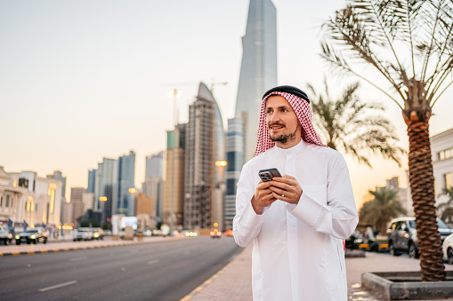 Handsome young man wearing keffiyeh and dishdasha and using his smart phone on the street in Kuwait city in Kuwait.