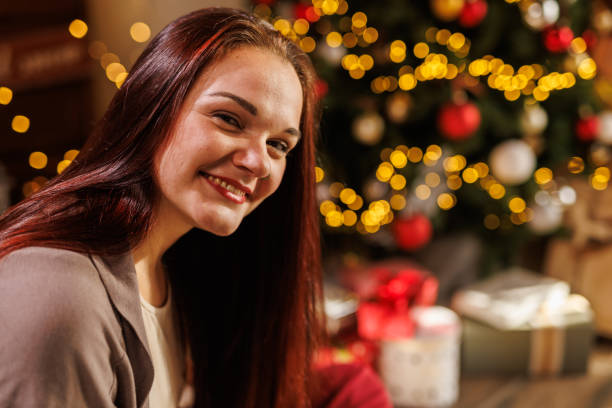 istock Young woman sitting by the Christmas tree, smiling joyfully at camera 2050227971