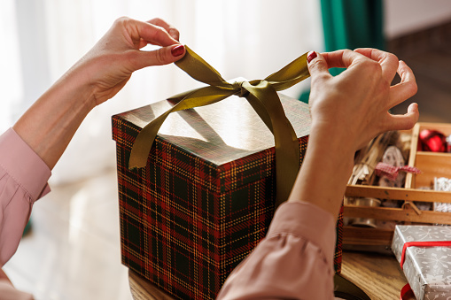 Close up shot of unrecognizable young woman tying a ribbon around a Christmas gift box that she is preparing for a loved one.