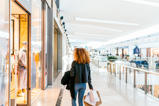 A young black girl with red afro hair walks through a shopping center looking at the windows of clothing stores, she carries shopping bags in her hand