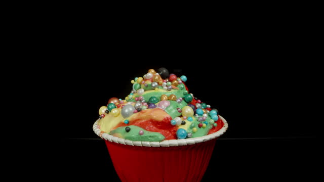 A cupcake with multicolored rainbow frosting and balls, isolated on a black background. Dolly slider, close up.