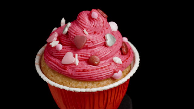 A woman's hand places a red heart on the pink cream of a cupcake for decoration.