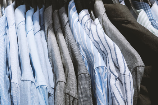 Multi-colored men's shirts hang in a row on hangers in store. Blue, light blue and gray shirts