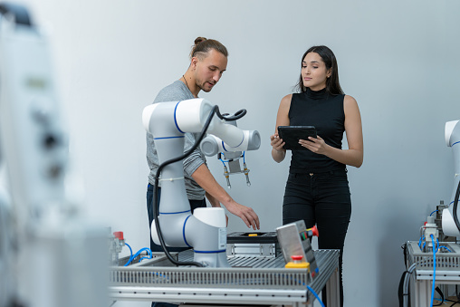 Latin American female engineer holding the controller to write program code to investigate automated robot arm while her handsome software engineer male coworker looking at the automated machine to inspect where the damage or error is at the innovation center.