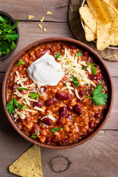 Chili con carne with cheese topings and nachos on wooden table, top view. Homemade mexican food, chili with red beans and ground beef. Chilean beans.