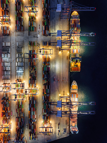 Aerial top down view of  large container cargo ship being unloaded by cranes and trucks in a commercial harbour during night time