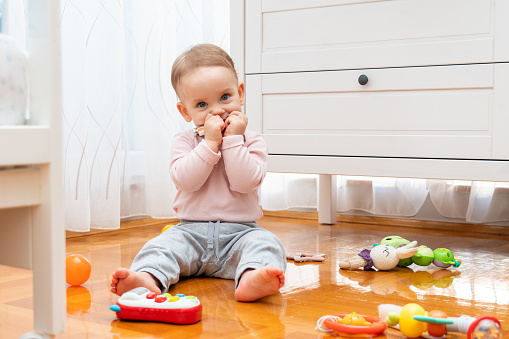 The baby hides his smile with his hands while sitting on the floor and playing with toys.