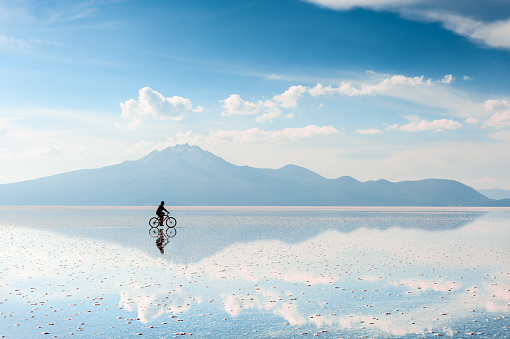 Uyuni, Bolivia - March, 26, 2017: Man tourist riding a bicycle on the Salar de Uyuni salt flat in Bolivia. Mountains and sky are reflected in the water surface