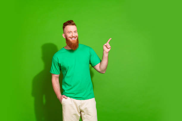 istock Photo of optimistic man with ginger beard dressed green t-shirt indicating at offer empty space isolated on green color background 2050550037