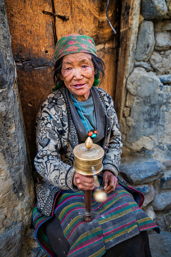 Old Tibetan woman sitting at the front of her house and praying in small village, Upper Mustang. Mustang region is the former Kingdom of Lo and now part of Nepal,  in the north-central part of that country, bordering the People's Republic of China on the Tibetan plateau between the Nepalese provinces of Dolpo and Manang.
