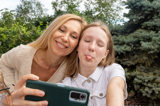 Girl shows tongue for goofy family selfie. Smiling woman and happy kid enjoy free time spending together in summer park cafe