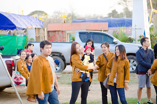 Thai people and family in classic brown jackets captured in Lampang. People are close to public market and festival around and below King Naresuan monument in Lampang. One woman has a baby on arms. A man is passing in foreground. Woman with baby is toothy smiling and looking. Other people sitting on a pick-up in background are smiling and looking too. At right side are some men.