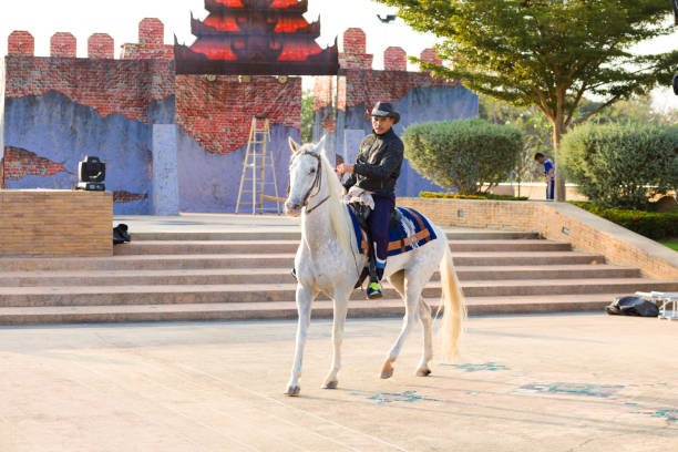 Thai man with cowboy hat on white horse is standing with horse on square close and below King Naresuan Monument in Lampang. In background are steps and trees and bushes. Some artificial historic looking wall decor is placed behind steps. Between trees a man is working. Monument was made for  history  of marching King Naresuan the Great Through Lampang. Monument was made in 1992 To honor Her Majesty His Majesty the Queen's fifth anniversary in 1992 for independence of the nation and place of worship for the general public and source of historical study