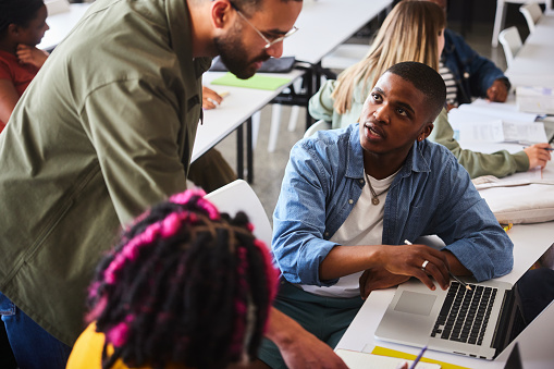 Young male college student asking his teacher a question while working on a  laptop during a lesson in a classroom at school