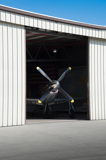 Partially opened aircraft hangar doors reveal four-blade propeller and glimpse of low wings and retractable landing gear of high performance airplane in a dark interior space. Dramatic.