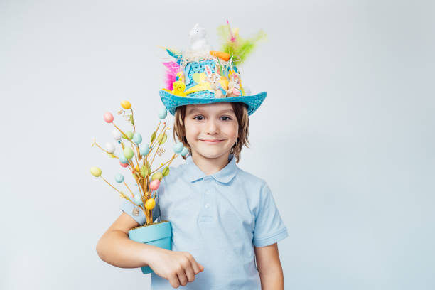 Easter eggs hunt tradition. Smiling boy kid in Easter hat holds easter tree in pot and chocolate egg in other hand on the white background. Easter decoration, family celebration, Christian traditions tradición de la caza de huevos de pascua. el niño sonriente con sombrero de pascua sostiene el árbol de pascua en la olla y el huevo de chocolate en la otra mano sobre el fondo blanco. decoración de pascua, celebración familiar, tradiciones cristianas
