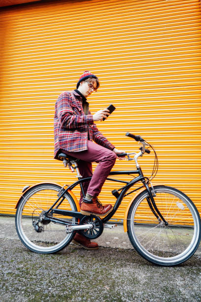 istock Young hipster man using mobile phone while sitting on city bicycle near orange wall. Student using 5g network,rests after riding. Eco friendly transport for carbon footprint. Sustainable lifestyle 2050645381
