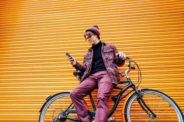 istock Young hipster man using mobile phone while sitting on cruiser bicycle near orange wall. Student using 5g network,rests after riding. Eco friendly transport for carbon footprint. Sustainable lifestyle 2050645390
