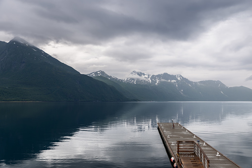 A wooden pier juts into the reflective fjord, drawing the eye towards the overcast Norwegian mountainscape, a serene yet moody atmosphere