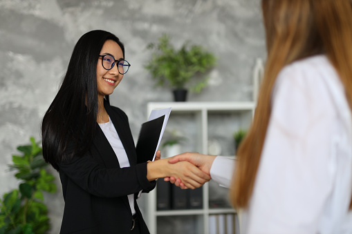 In a corporate office, an Asian businesswoman and caucasian businesswoman seal a successful partnership with a handshake.