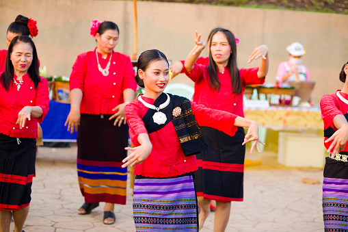 Group of rum thai dancing adult thai women in traditional clothing with focus of woman in center and foreground. Scene is at a market and local festical below King Naresuan monument in Lampang. Women are cheerful and smiling. Women are wearing red shirts