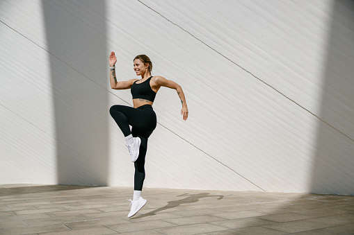 Young fit woman in sportswear leaping in the air with wall backdrop outdoors. High quality photo