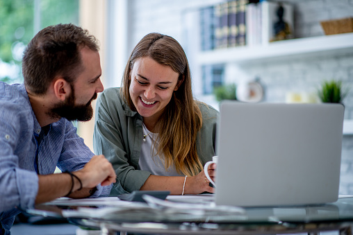 A young couple sit together in their home office as they work through paying bills together.  They are both dressed casually, have a laptop open in front of them and appear happy.