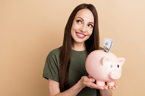 Portrait of dreamy mature woman hold piggy bank looking curious novelty puts banknote inside box isolated on beige color background.