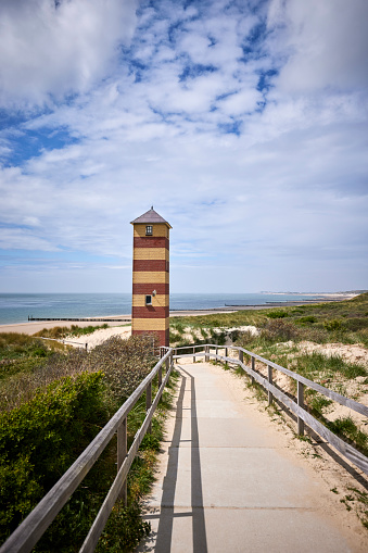 Lighthouse of Dishoek along the Dutch coast during a sunny day. There is a path to the beach with a fence, the lighthouse is red and yellow striped.