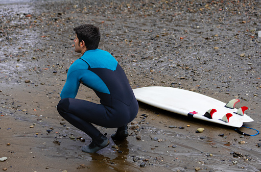 A surfer crouches thoughtfully on a rocky beach next to his surfboard, contemplating the ocean before surfing.