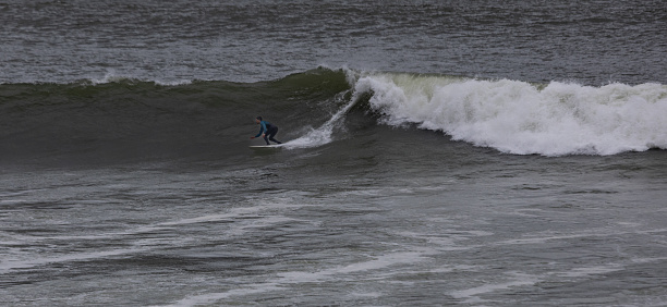 A solitary surfer rides a wave on a grey, overcast day, showcasing the sport's thrill under dramatic weather conditions.